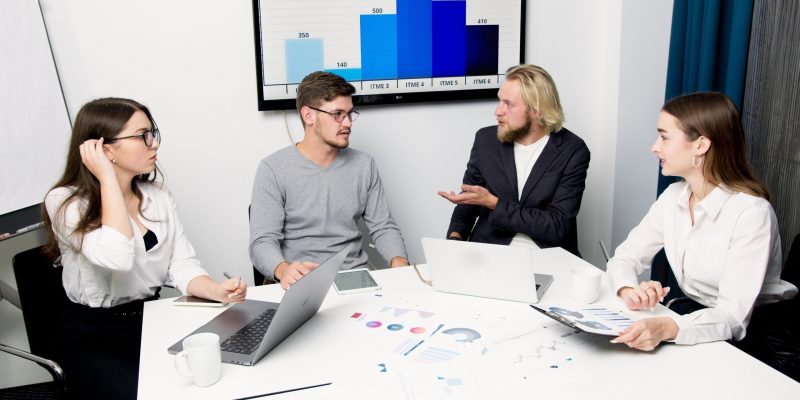 Four professionals collaborating during a People Analytics Consulting Services session, discussing workforce insights around a table with charts, documents, and graphs displayed in the background to support data-driven organizational planning and performance analysis.